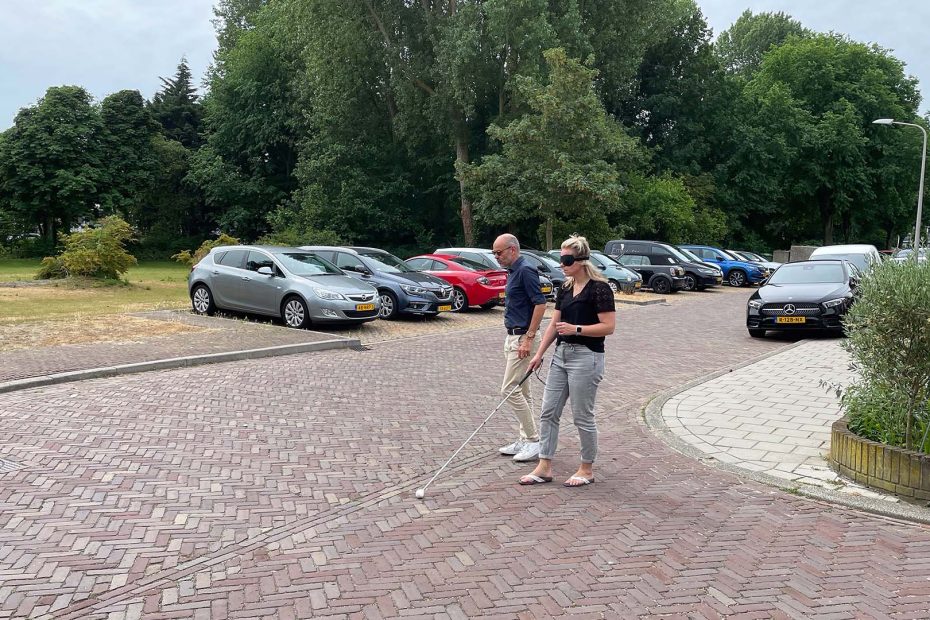 Een vrouw met een slecht zicht bril en een witte taststok steekt de straat over. Ze wordt begeleid door een man die naast haar loopt.